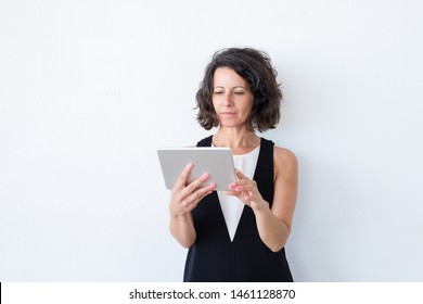 Serious Focused Woman In Casual Using Tablet. Curly Haired Middle Aged Model Standing Over White Background And Reading On Screen. Tablet Using Concept