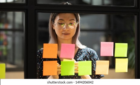 Serious focused young female coach teacher student asian business woman working on project strategy plan writing target tasks creative ideas on sticky post it notes on glass scrum board office wall