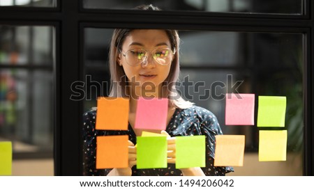 Serious focused young female coach teacher student asian business woman working on project strategy plan writing target tasks creative ideas on sticky post it notes on glass scrum board office wall