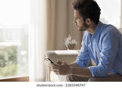 Serious freelance business man using application on smartphone for online communication, sitting in city home apartment, holding gadget, looking at window away, thinking, making decision - Shutterstock ID 2283348747
