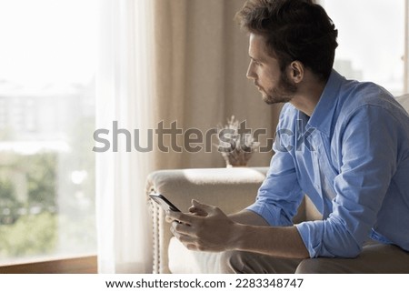 Serious freelance business man using application on smartphone for online communication, sitting in city home apartment, holding gadget, looking at window away, thinking, making decision