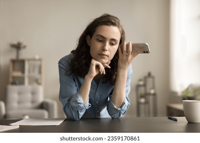 Serious freelancer employee girl holding mobile phone at head, listening to sound, voice message, audio file, leaning hands on home workplace table, thinking, touching chin - Powered by Shutterstock