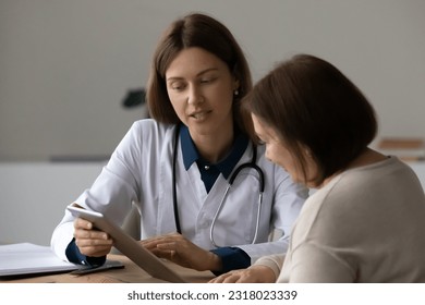 Serious friendly doctor talking to senior mature patient, using gadget, showing tablet screen, telling about treatment, explaining examination result, giving consultation at home or in hospital office - Powered by Shutterstock