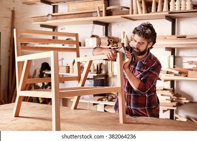 Serious furniture designer carefully sanding a chair frame that he is busy manufacturing in his woodwork studio, with shelves of wooden items behind him