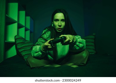Serious gamer girl plays games on console with gamepad in hands at home in green light, focused looking at camera, close-up portrait.