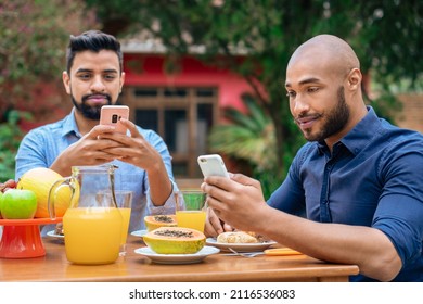 Serious Gay Couple Using Mobile And Having Breakfast Outside In The Garden