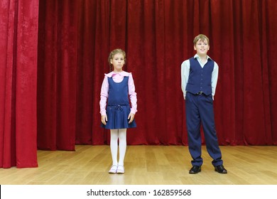 Serious Girl And Happy Boy In School Uniform Stand On Stage With Red Curtains.