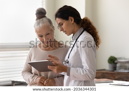 Serious GP doctor showing tablet screen to old 70s female patient, explaining electronic prescription, medical screening, examination result, giving consultation. Woman visiting practitioner