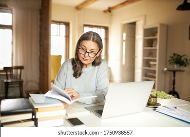 Serious Gray Haired Female Teacher In Her Sixties Sitting At Desk Surrounded With Textbooks, Making Lesson Plan Using Laptop. Middle Aged Woman Working Distantly From Home, Holding Open Book