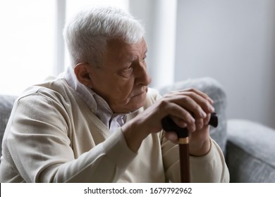 Serious Gray-haired Man Hold Wooden Walking Stick Sit On Couch In Living Room Alone Look At Distance Feels Upset And Lonely. Nursing Home For Older Disabled People, Senile Diseases, Healthcare Concept