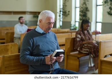 Serious Grey Haired Man In Casualwear Holding Bible And Rosary Beads In Front Of Himself During Silent Pray Against Black Woman In Church