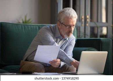 Serious Grey Haired Mature Man In Glasses Calculating Bills, Using Laptop, Online Bank Service At Home, Focused Middle Aged Grandfather Looking At Computer Screen, Typing, Checking Loan Documents