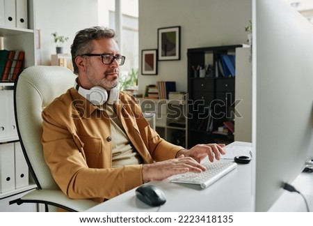 Serious grey haired programmer looking at computer screen while sitting by workplace in front of monitor and decoding data