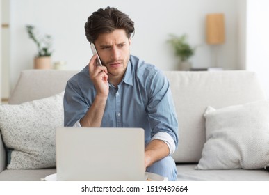 Serious Guy Having Unpleasant Phone Conversation Working On Laptop Sitting On Sofa At Home. Empty Space