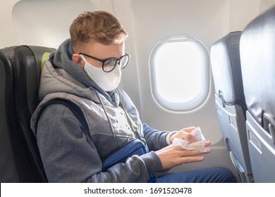 Serious Guy, Young Man On Airplane, Plane In Glasses And Medical Protective Sterile Mask On His Face Disinfect, Clean Hands With Napkin, Traveling. Coronavirus, Virus Concept. Pandemic Covid-19.