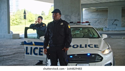 Serious Handsome African American Police Officer Looking Around Standing By Patrol Car. Two Young Policemen On Duty Keep Guard On City At Daytime.