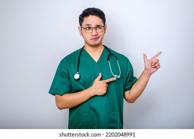 Serious Handsome Asian Doctor, Nurse In Scrubs And Stethoscope, Pointing Fingers Upper Right Corner, Showing Online Clinic Banner, White Background