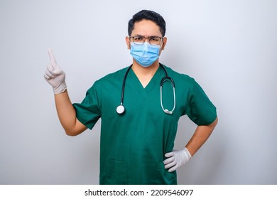 Serious Handsome Asian Doctor, Nurse In Scrubs And Stethoscope, Pointing Fingers Upper Left Corner, Showing Online Clinic Banner, White Background