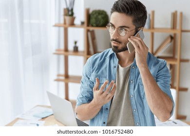 Serious Handsome Bearded Male Company Owner Has Phone Conversation, Manages Finances On Distance, Gestures As Tries To Explain Something To His Interlocutor, Stands Against Office Interior Indoor