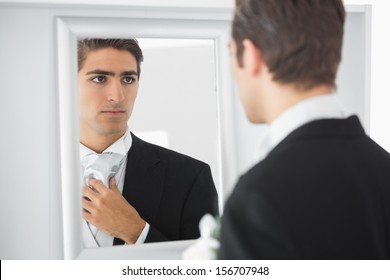 Serious handsome bridegroom looking in mirror straightening his tie 