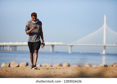 Serious Handsome Young Black Guy With Headphones On Neck Texting Sms While Walking Over Beach In Summer