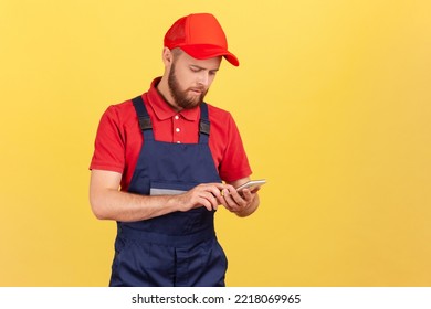 Serious Handyman In Overalls Typing On Mobile Phone, Using Cellphone Messenger To Accept Online Order As Delivery, Repair And Maintenance Services. Indoor Studio Shot Isolated On Yellow Background.