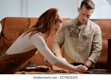 serious hardworking girl concentrated on cutting leather at workshop. handsome working guy in the blurred background - Powered by Shutterstock