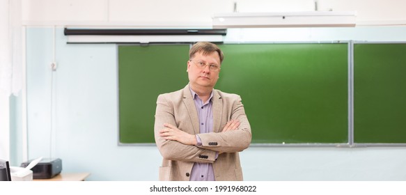 The serious headmaster stands in the classroom at the blackboard with his arms folded. - Shutterstock ID 1991968022