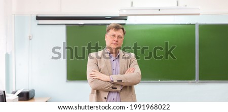 The serious headmaster stands in the classroom at the blackboard with his arms folded.