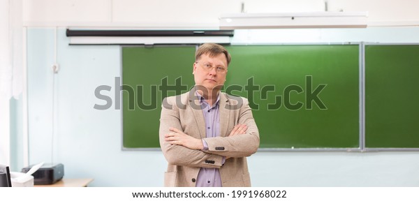 The serious headmaster stands in the\
classroom at the blackboard with his arms\
folded.