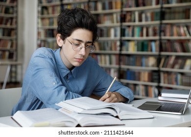 Serious high school pupil working on graduation project in library. Focused college student guy in glasses writing notes, lesson summary at laptop, watching webinar, reading open books