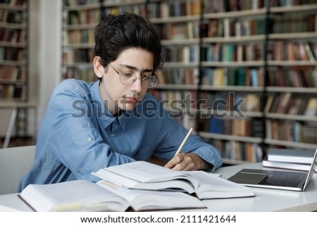 Serious high school pupil working on graduation project in library. Focused college student guy in glasses writing notes, lesson summary at laptop, watching webinar, reading open books