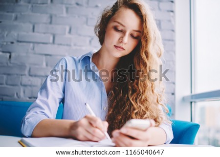 Serious hipster girl spending time for writing organisation plan in textbook while making research on smartphone device using free 4g internet, attractive female student doing school homework