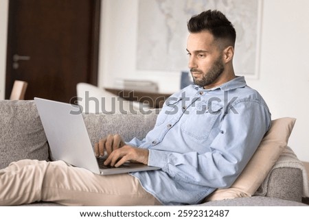 Serious Hispanic man settled down comfortable on sofa with wireless computer, typing, teleworking, choosing goods, booking hotel, buy tickets, studying, gains new knowledge through new modern platform
