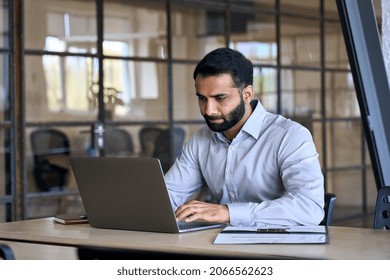 Serious Indian Business Man Employee Professional Manager Using Computer, Watching Webinar Working In Modern Office Doing Online Data Market Analysis Thinking Planning Tech Strategy Looking At Laptop.