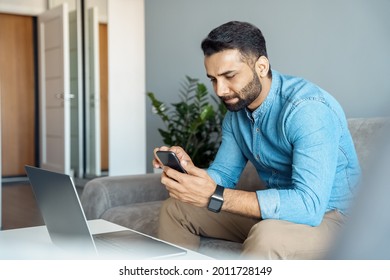 Serious Indian Business Man Using Smartphone. Eastern Entrepreneur Making Online Video Call On Mobile Phone While Working Laptop Computer With Internet Technology Sitting On Sofa At Home Office