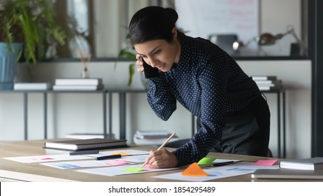 Serious Indian businesswoman talking on cellphone, writing notes on colorful sticky papers in office, employee checking financial documents, working with statistics, consulting client by phone call
