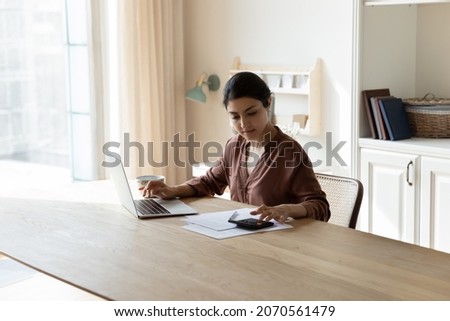 Serious Indian housewife sit at table busy in personal expenses management, use calculator calculate household utilities, pay bills through secure e-banking app on laptop. Finances, accounting concept