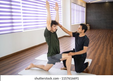 Serious Indian Instructor Helping Beginner At Yoga Class. Man Doing Warrior Pose In Gym. Personal Yoga Instructor Concept.