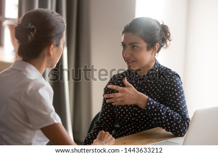 Serious indian mentor teacher worker talk to female colleague teach intern discussing new skills learning sit at work desk, two diverse coworkers work together help cooperate on project in teamwork
