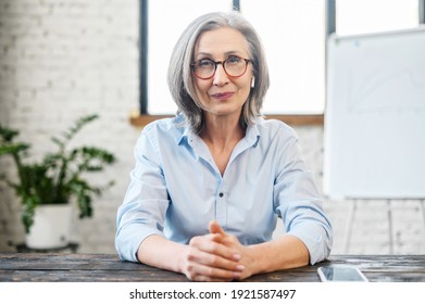 Serious intelligent mature senior teacher businesswoman with pleasant smile and earphones in glasses looking at camera, sitting at the desk, offering professional services, help, feeling ready to work - Shutterstock ID 1921587497