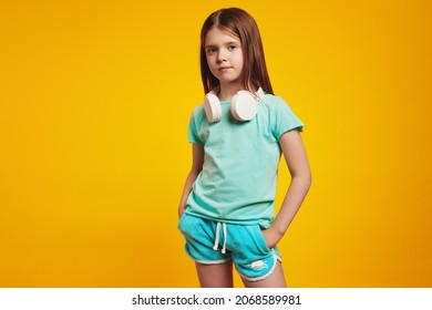 Serious Kid Girl In Stylish Summer Outfit, Wearing White Headphones On Neck, Keeping Hands In Pockets, Isolated Over Yellow Brick Wall