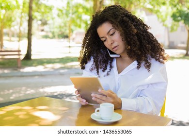 Serious lady calling on phone and using tablet in outdoor cafe. Beautiful woman wearing blouse and sitting with blurred green trees in background. Leisure and communication concept. Front view. - Shutterstock ID 1436553065