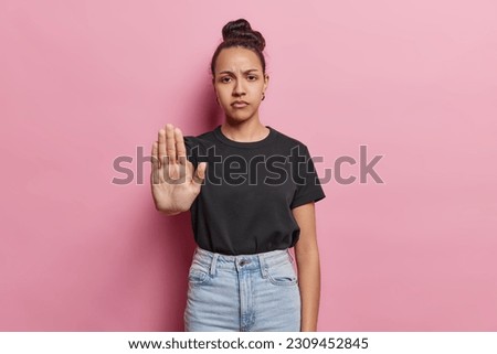 Serious Latin woman shows stop gesture with palm asserts boundaries sends message of resistance has strict expression wears black t shirt and jeans poses against pink background. Self empowerment