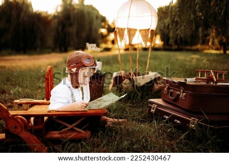 Serious little pilot boy in vintage aviator hat sitting and studying a map near wooden plane outdoor. Happy childhood. Dreams come true. 