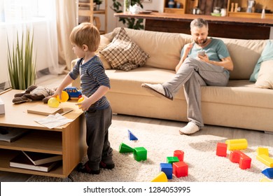 Serious Little Son In Casual Clothing Standing At Coffee Table And Picking Up Toys While Father Using Gadget And Sitting On Sofa