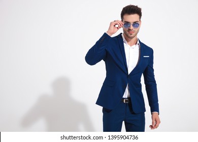 Serious looking guy adjusting his sunglasses while wearing a blue suit and standing on white studio background