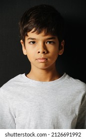 Serious Looking Indian Boy In A White T Shirt Over Black Background. Standing Straight, Looking At The Camera.