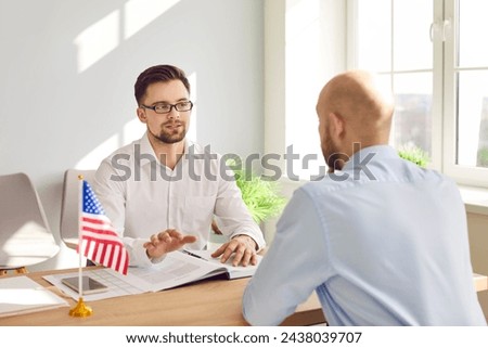 Serious male Consul of United States of America sitting at office table with American flag and documents and talking to young man about his immigration plans and USA travel visa application