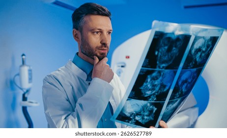 Médico masculino serio estudiando radiografía craneal con la mano apoyada en la barbilla. Sostener una película de exploración detallada mientras se revisan las Imágenes de diagnóstico junto al escáner de TC. Concentrándose en el entorno médico moderno.